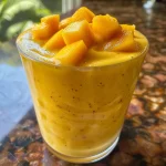 Batido de mango fresco servido en un vaso transparente con ingredientes alrededor.