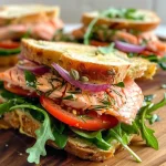 ['Sándwich de salmón con hierbas y aderezo de limón y alcaparras.', 'Primer plano de un sándwich de salmón fresco con vegetales.', 'Delicioso sándwich de salmón con condimentos y vegetales frescos.', 'Sándwich de salmón con hierbas, mayonesa y verduras en pan.']